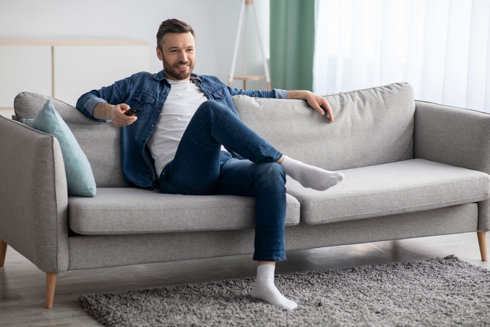 Watching TV at home. Relaxed middle-aged man having rest, switching channels with remote controller and smiling, empty space. Bearded man enjoying his weekend, watching TV in living room